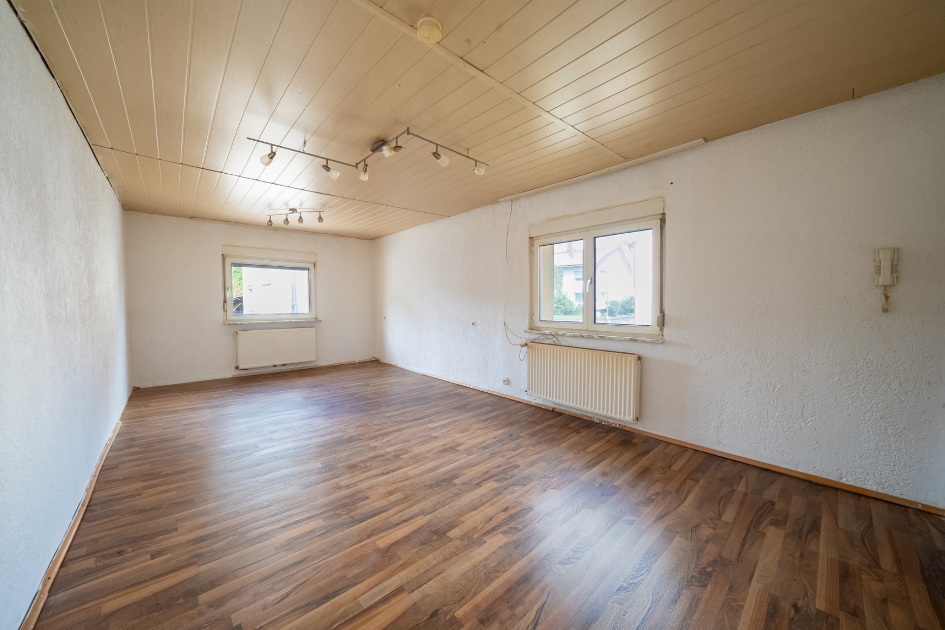 Leerer Raum mit Holzfußboden, zwei Fenstern, weißen Wänden und Deckenleuchten.