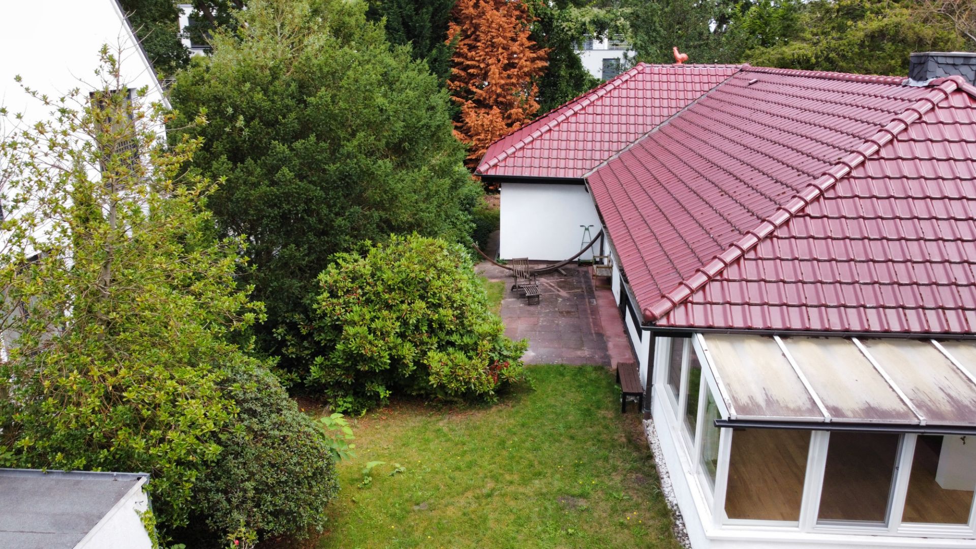 Garten mit Rasenfläche, Büschen und einem Haus mit rotem Dach und Wintergarten.