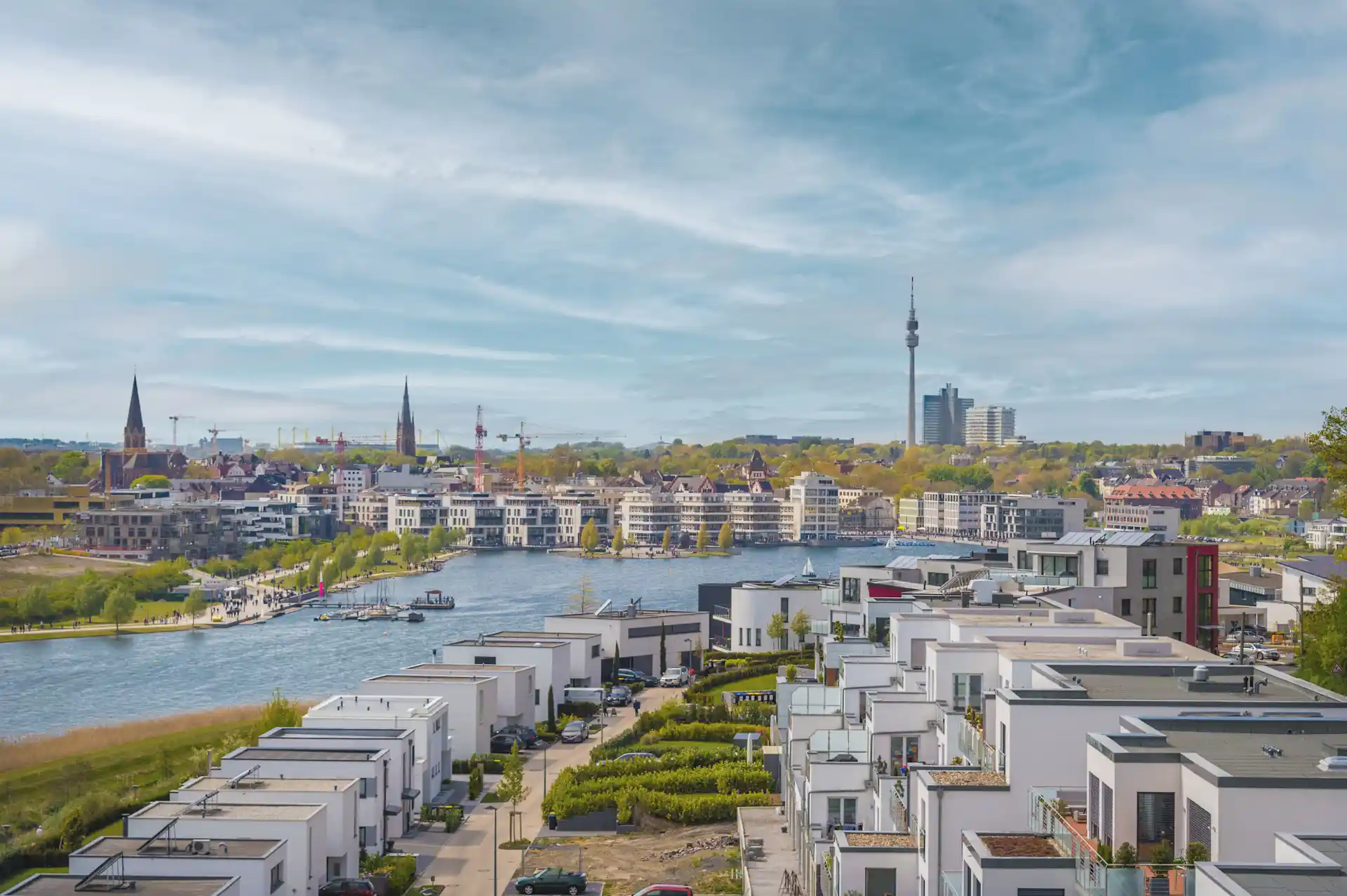 Moderne Wohnhäuser an einem Fluss, im Hintergrund Stadt mit Kirchtürmen und Fernsehturm.