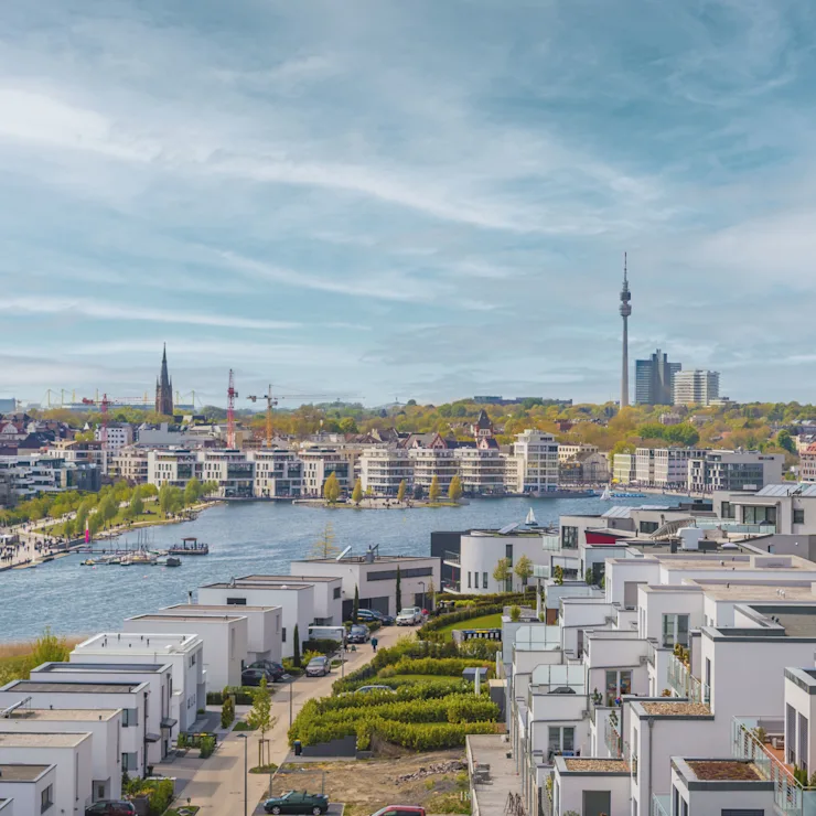 Moderne Wohnhäuser an einem Fluss, im Hintergrund Stadt mit Kirchtürmen und Fernsehturm.