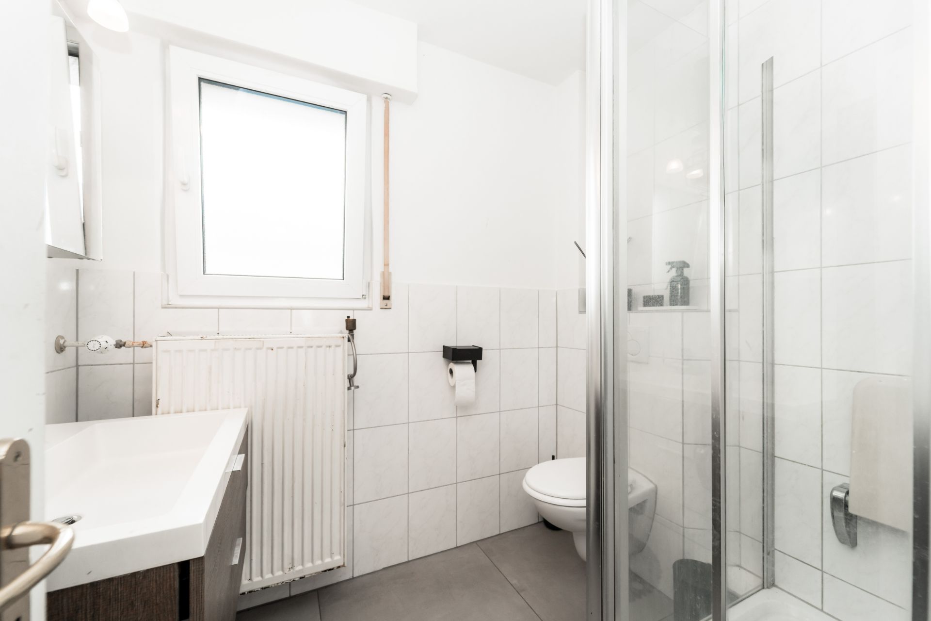 Helles Badezimmer mit Fenster, Waschbecken, Toilette, Heizung und Dusche mit Glastür.
