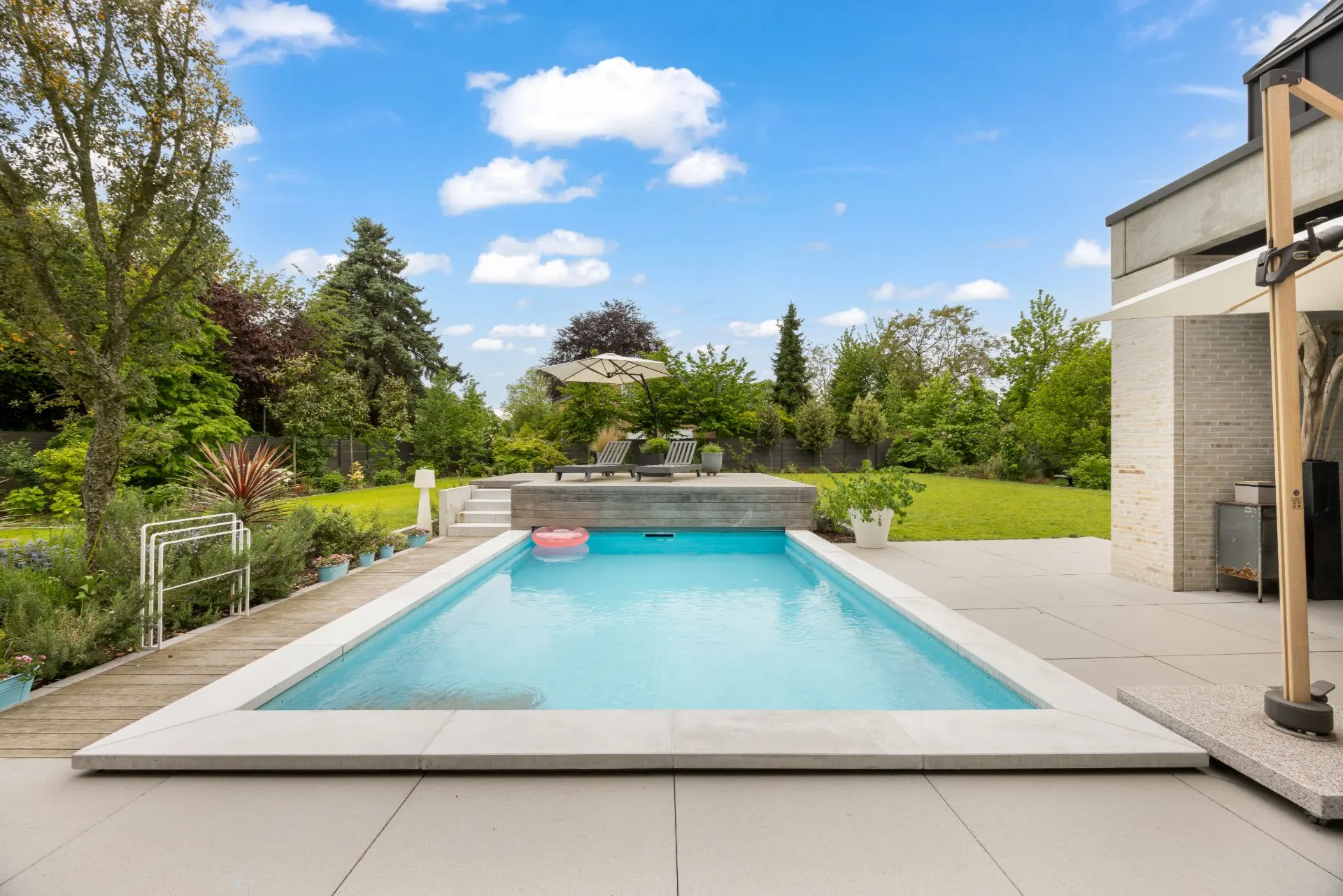 Moderner Garten mit Pool, Terrasse und Liegen, umgeben von Bäumen und grüner Wiese.