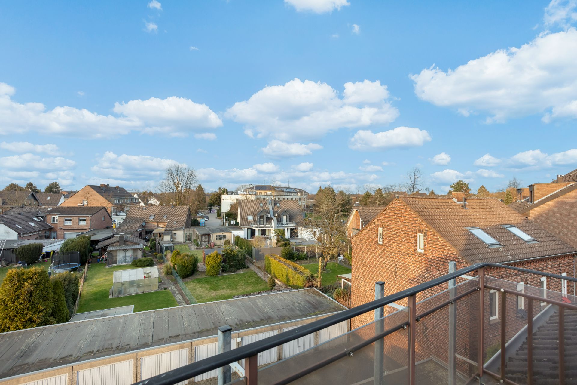 Blick von einem Balkon auf Nachbarhäuser, Gärten und einen blauen Himmel mit Wolken.