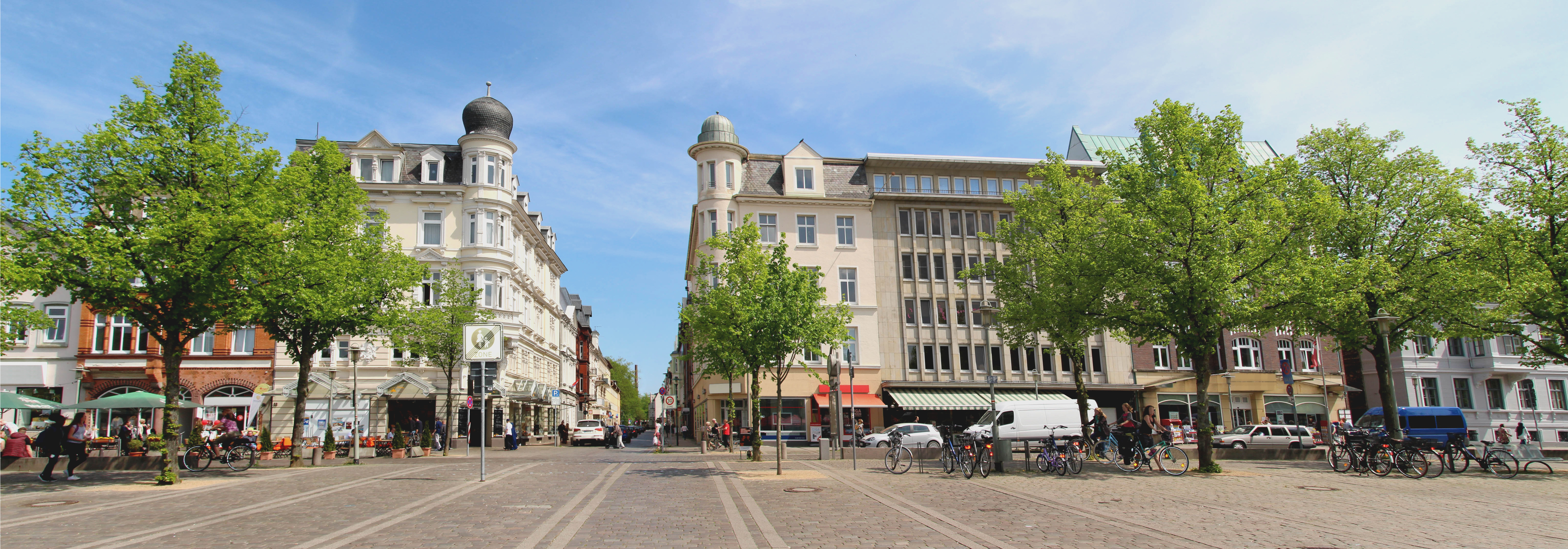 Städtischer Platz mit Bäumen, mehreren Wohngebäuden und geparkten Fahrrädern.