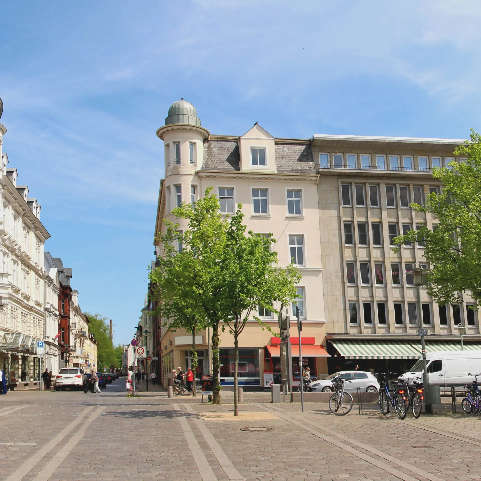 Städtischer Platz mit Bäumen, mehreren Wohngebäuden und geparkten Fahrrädern.