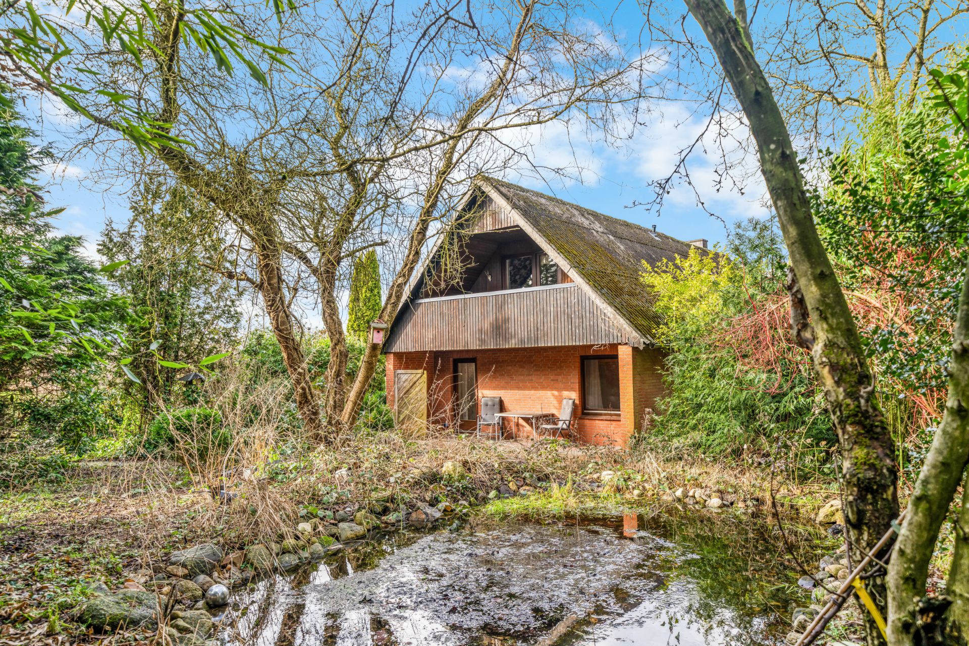 Backsteinhaus mit Balkon und kleinem Teich im zugewachsenen Garten, umgeben von Bäumen.