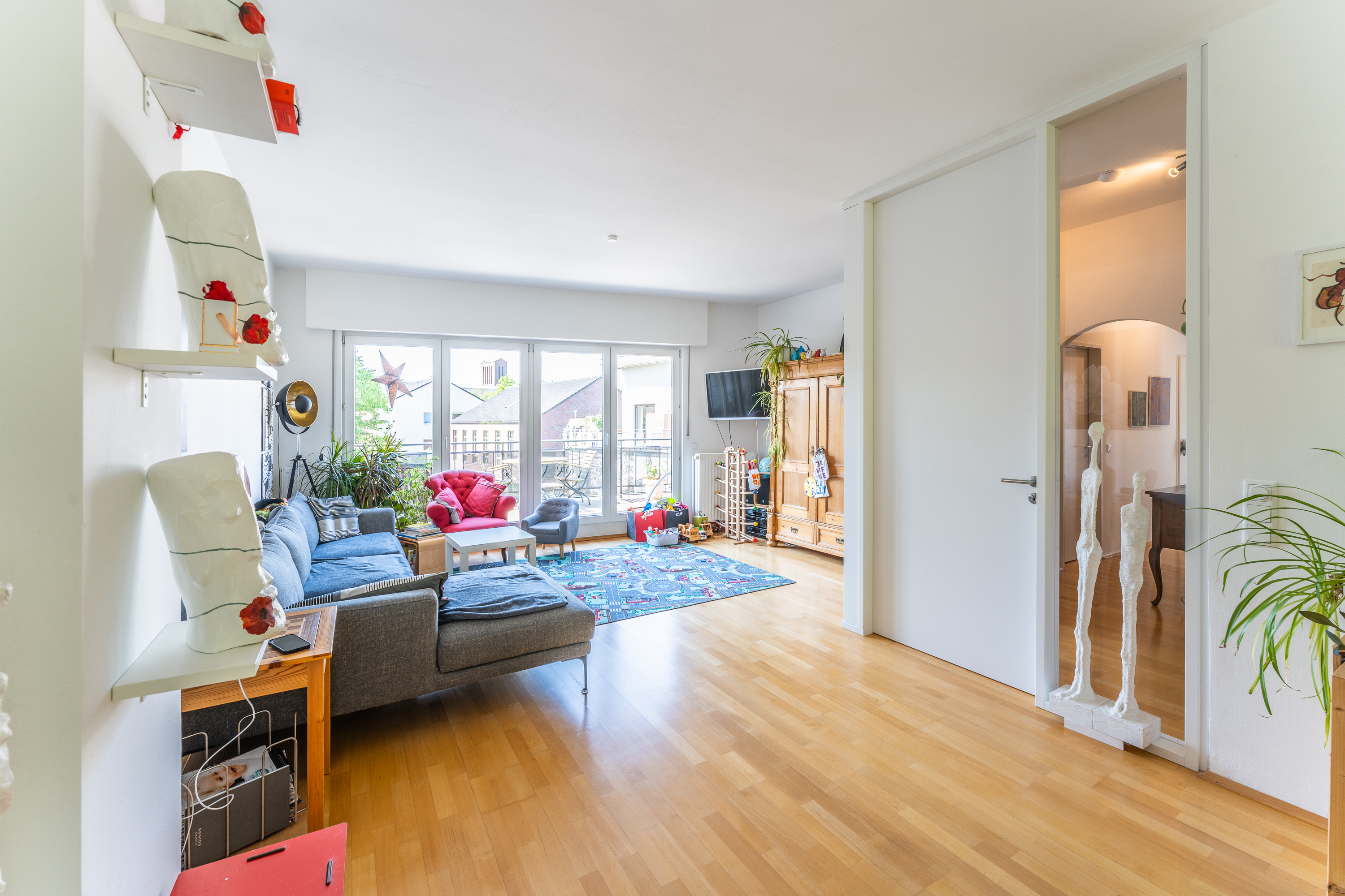 Helles Wohnzimmer mit grauem Sofa, Holzschrank, Pflanzen und großem Fenster zur Terrasse.