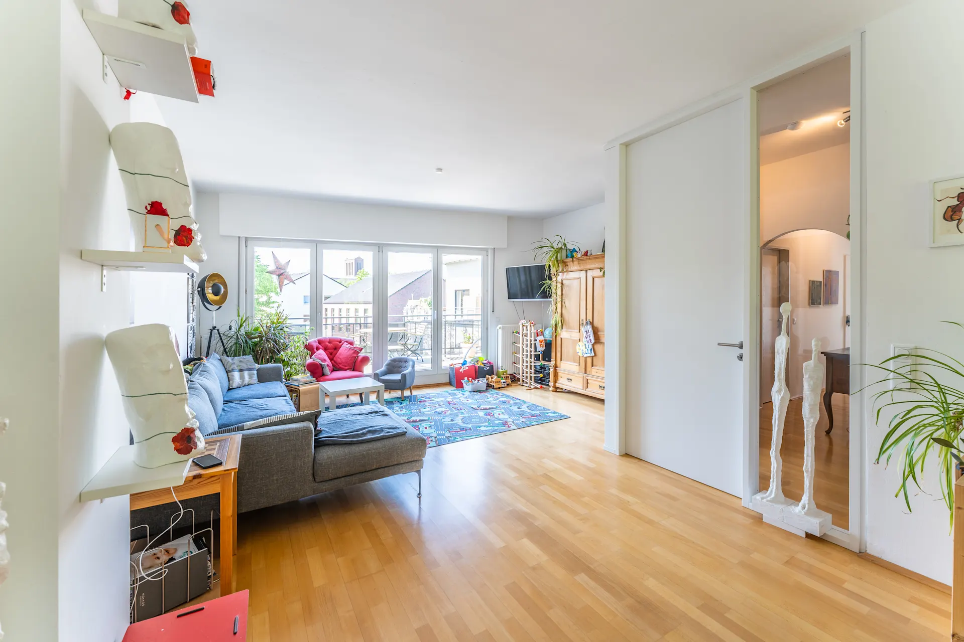 Helles Wohnzimmer mit grauem Sofa, Holzschrank, Pflanzen und großem Fenster zur Terrasse.