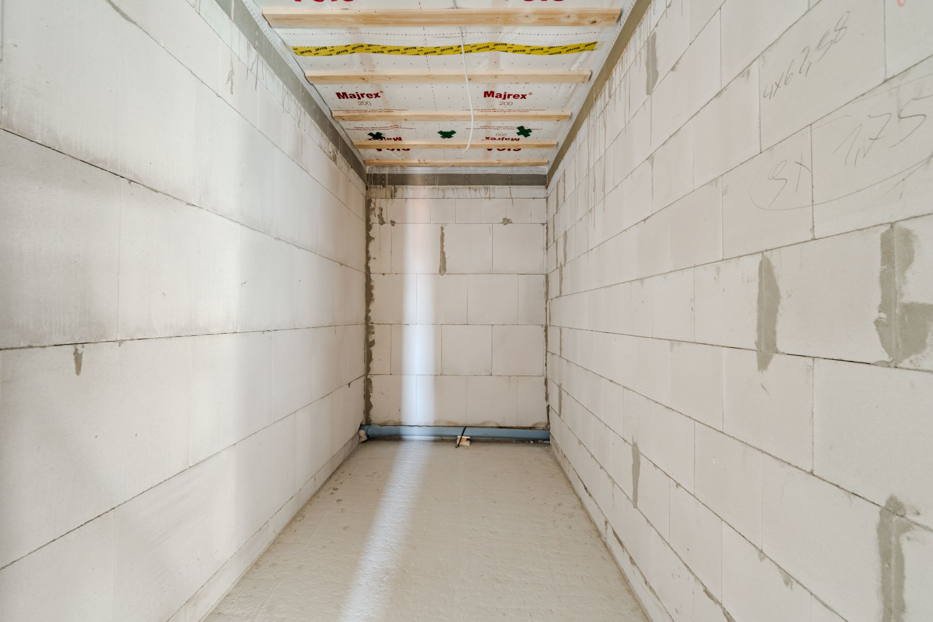 Schmaler Raum im Rohbau mit weißen Wänden und sichtbaren Leitungen am Boden.
