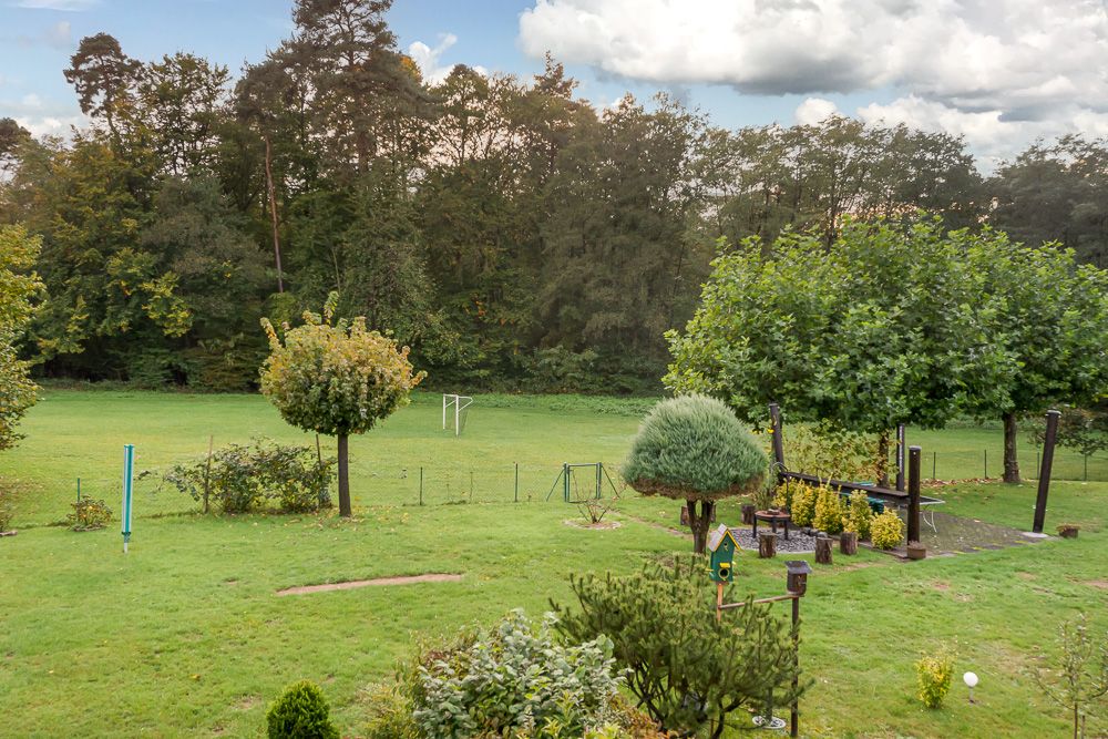 Großer Garten mit Rasen, Bäumen, Sitzbereich und Spielplatz, angrenzend an ein Waldstück.