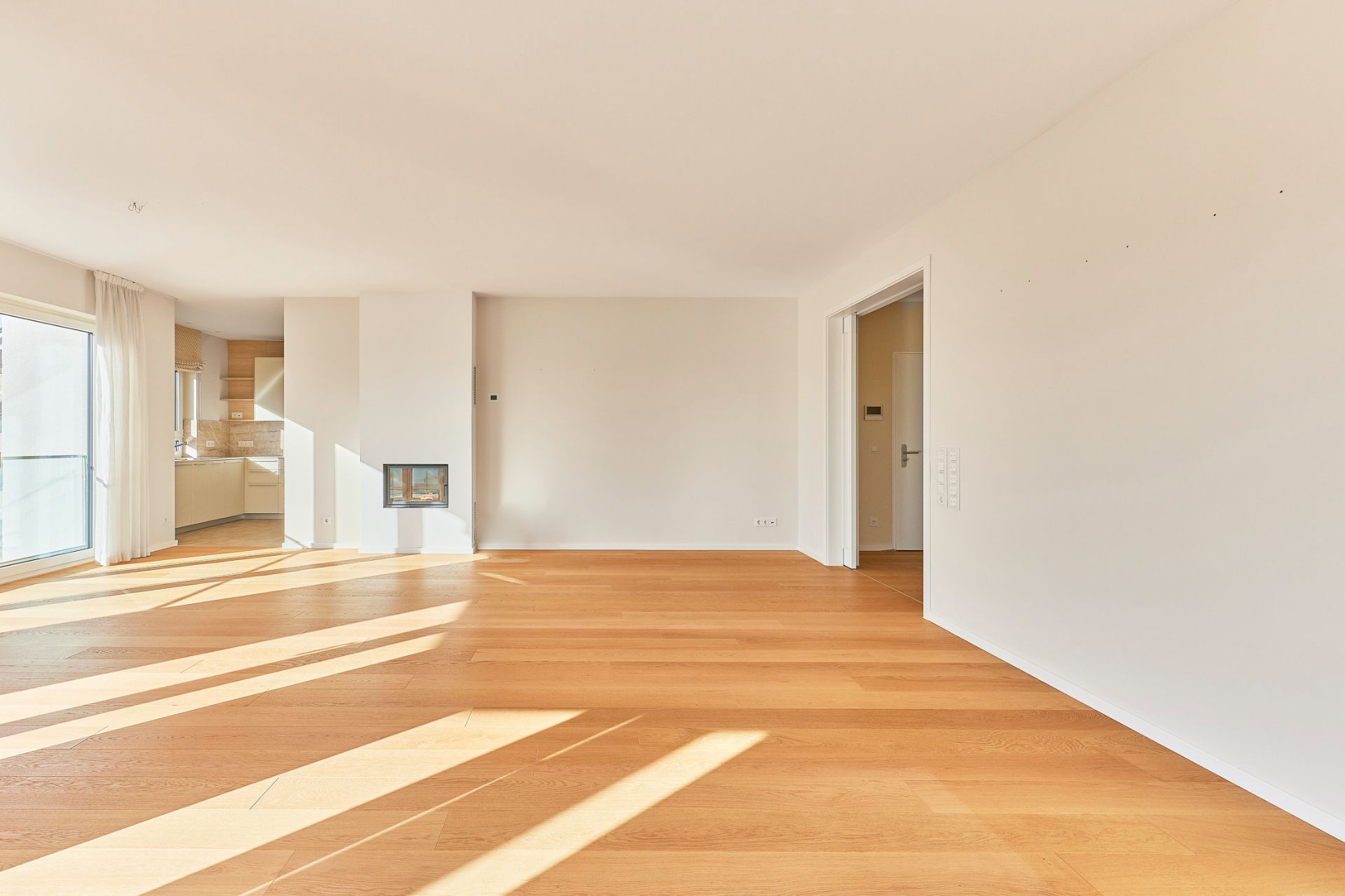Heller Wohnraum mit Holzfußboden, großen Fenstern und Zugang zur offenen Küche.