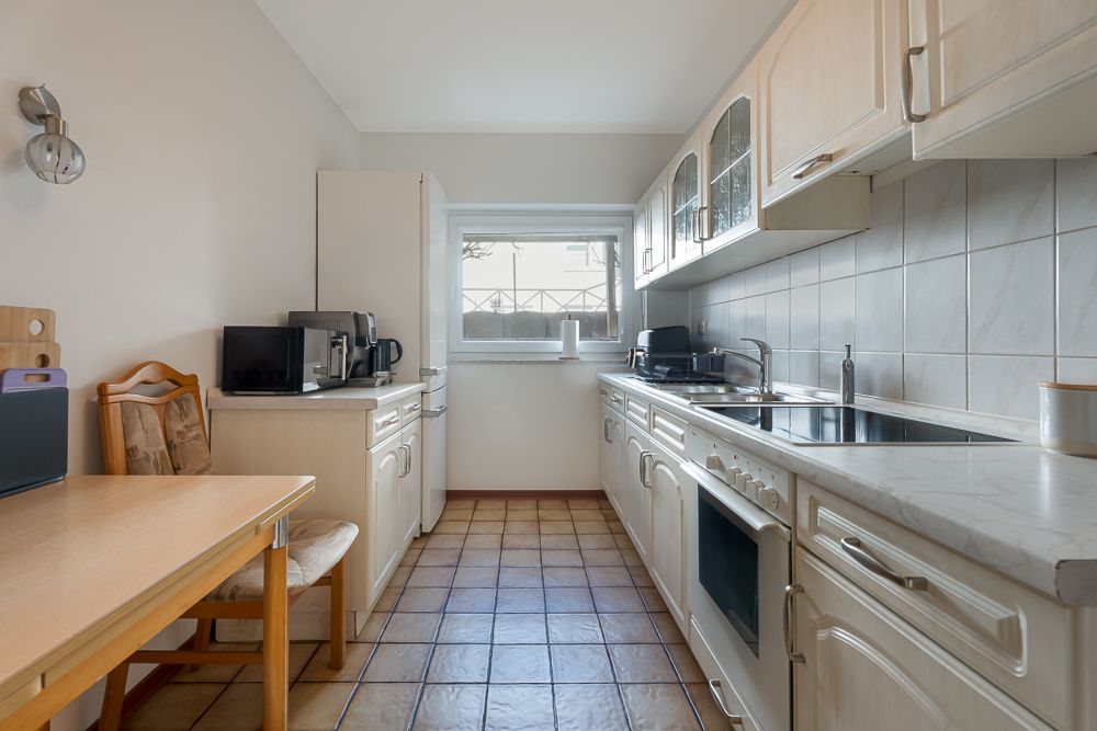 Helle Küche mit Holzmöbeln, Elektrogeräten, Tisch und Stuhl, Fliesenboden und Fenster.