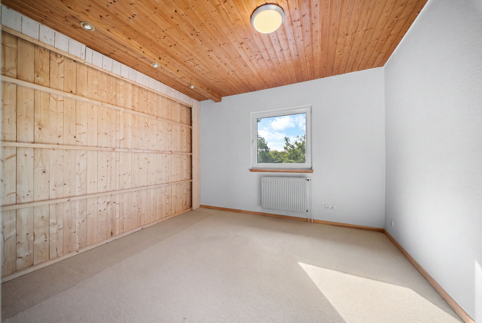 Helles, leeres Zimmer mit Holzdecke, Holzregalwand und Fenster mit Blick nach draußen.