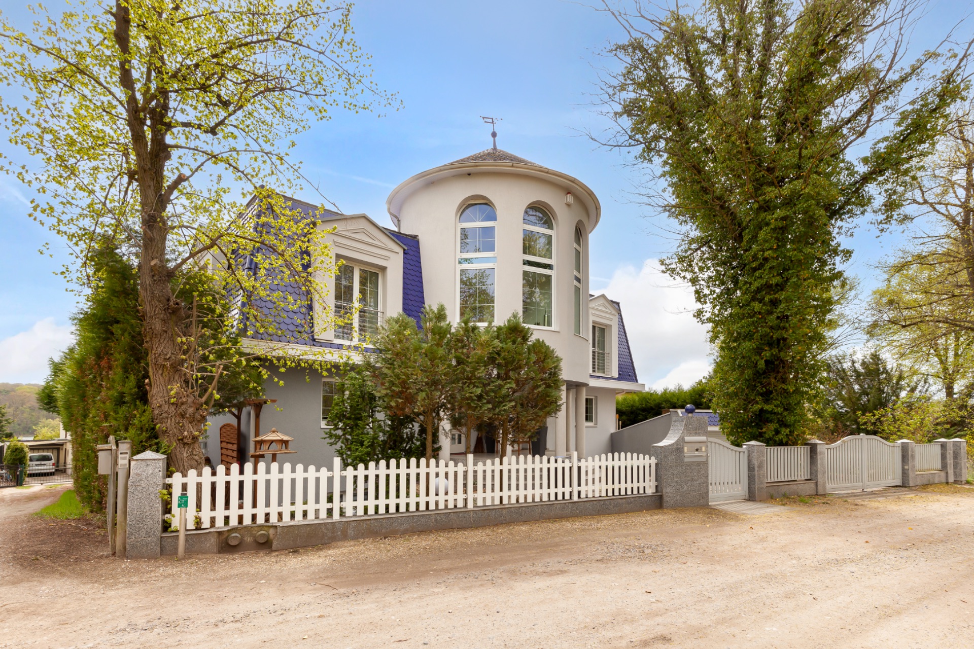 Großes Wohnhaus mit rundem Turm, weißen Fenstern und einem weißen Gartenzaun vor Bäumen.