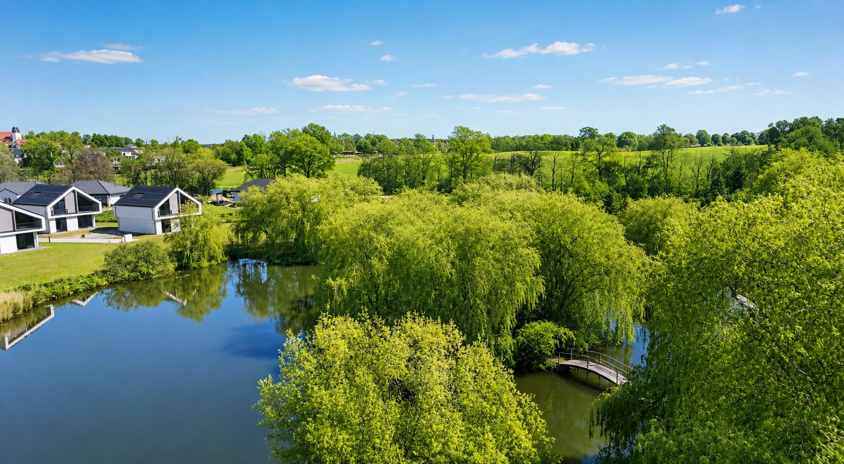 Teich mit Brücke, umgeben von Bäumen, moderne Wohnhäuser im Hintergrund bei sonnigem Wetter.
