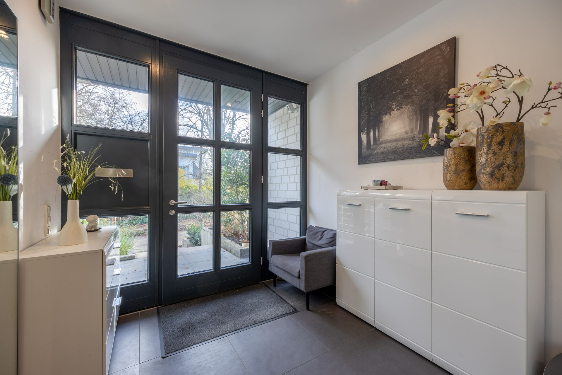 Heller Flur mit Glastür, weißem Schrank, Sessel und Blumenvasen.