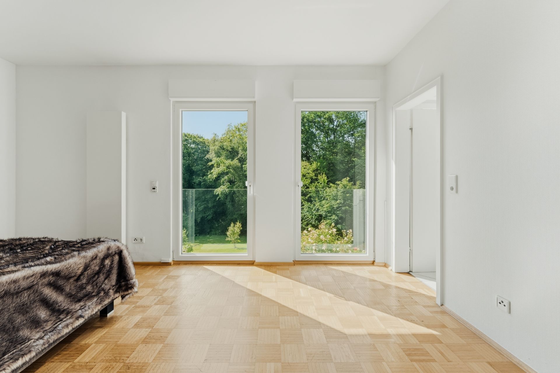 Helles Schlafzimmer mit Parkettboden, Bett links und zwei großen Fenstern mit Blick ins Grüne.