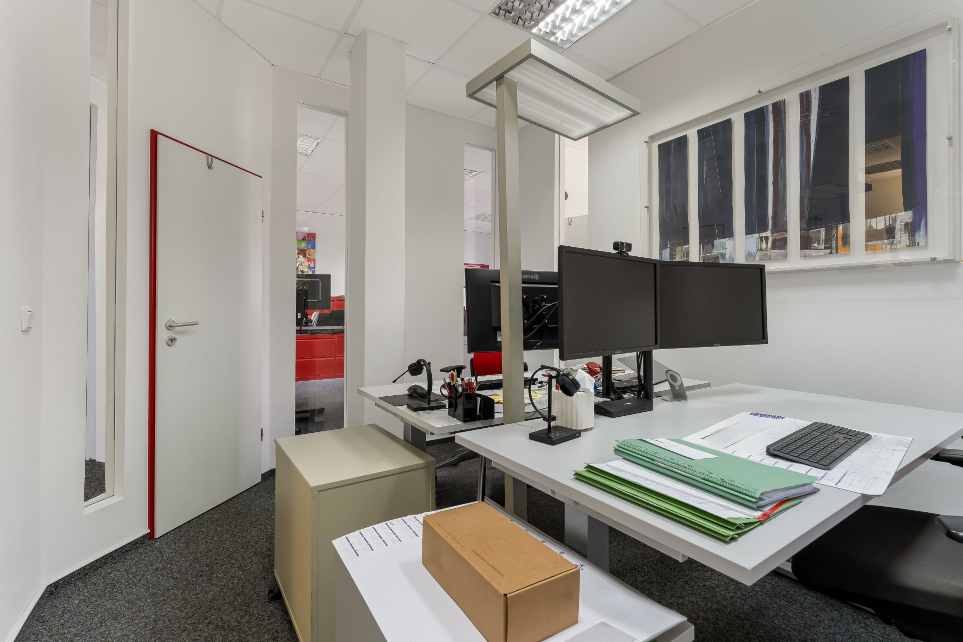Modernes Arbeitszimmer mit zwei Monitoren, Schreibtisch, Papieren und Lampe in heller Umgebung.