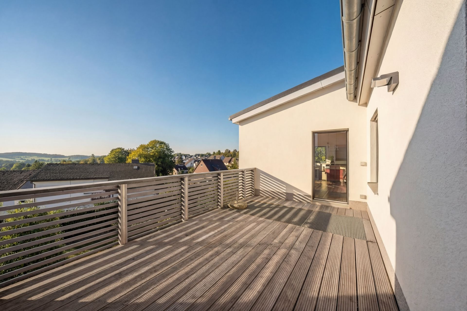 Große Holzterrasse mit Geländer, Blick auf Nachbarhäuser und Bäume bei klarem Himmel.