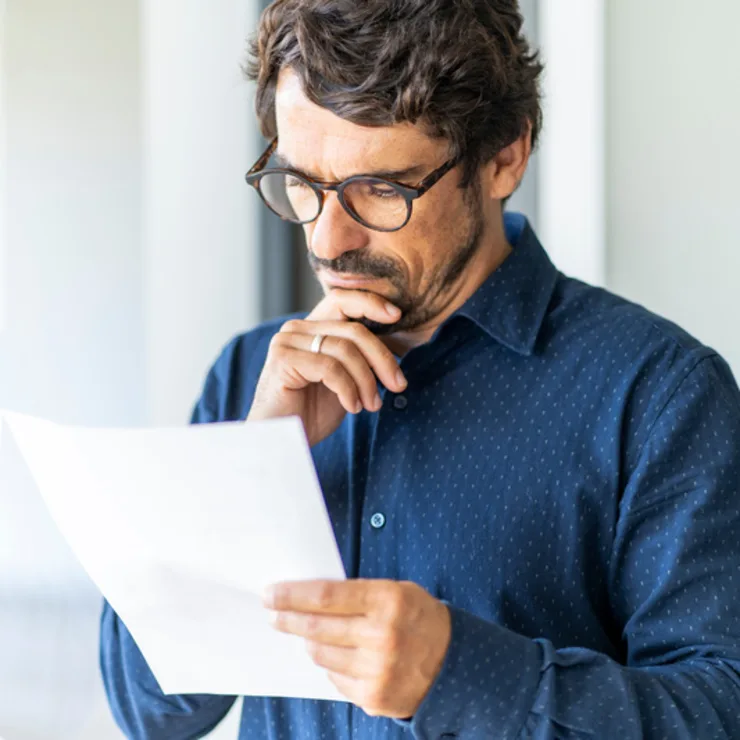 Mann mit Brille liest nachdenklich ein Blatt Papier in einem hellen Raum.
