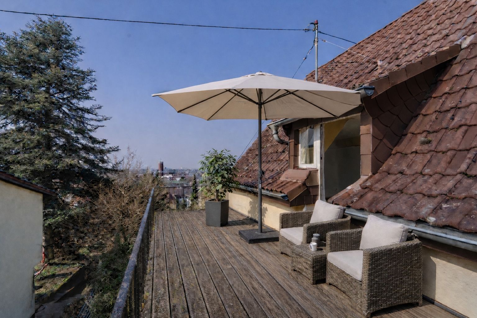 Holzterrasse mit zwei Stühlen, Sonnenschirm und Pflanze vor einem Haus mit Ziegeldach.