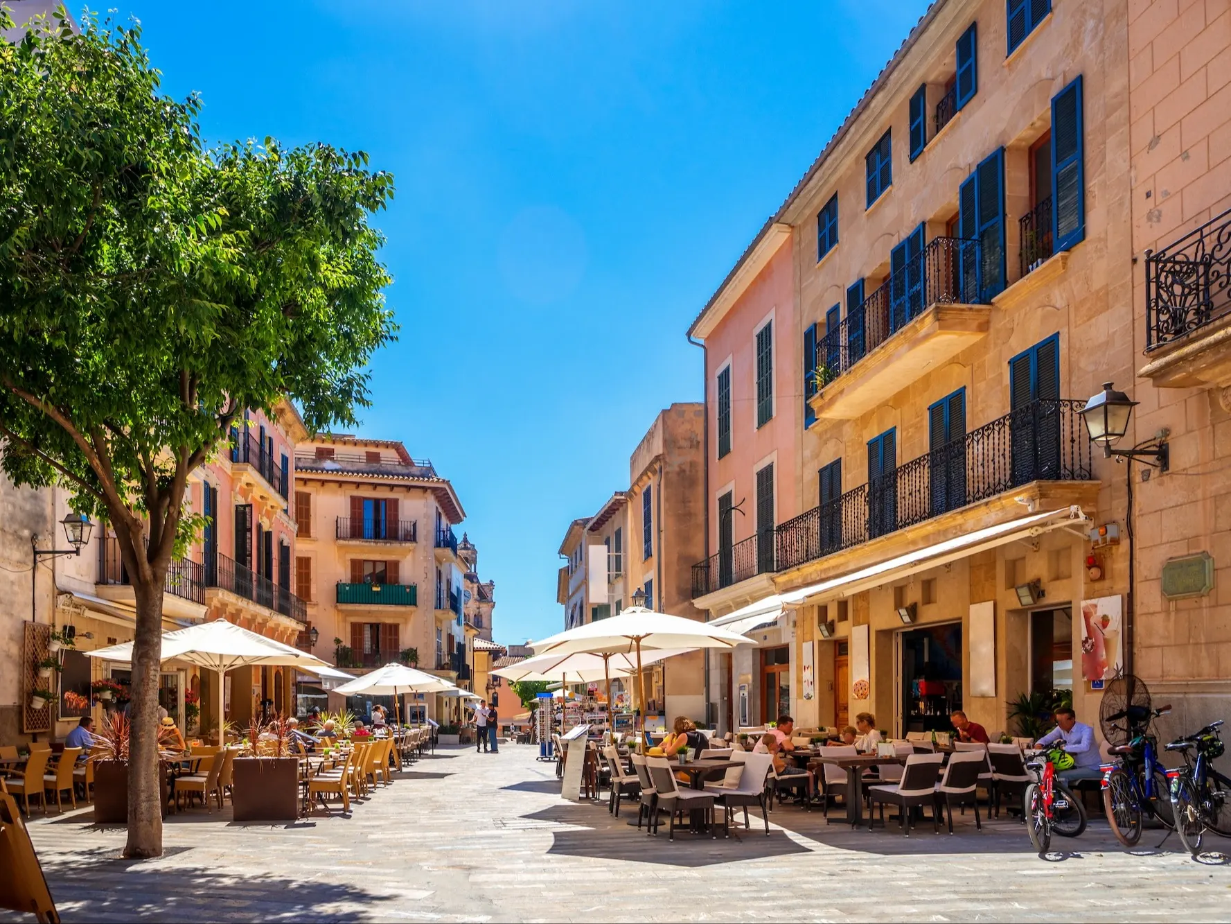 Sonnige Straße mit Wohnhäusern, Bäumen und Straßencafés mit Menschen unter Sonnenschirmen.