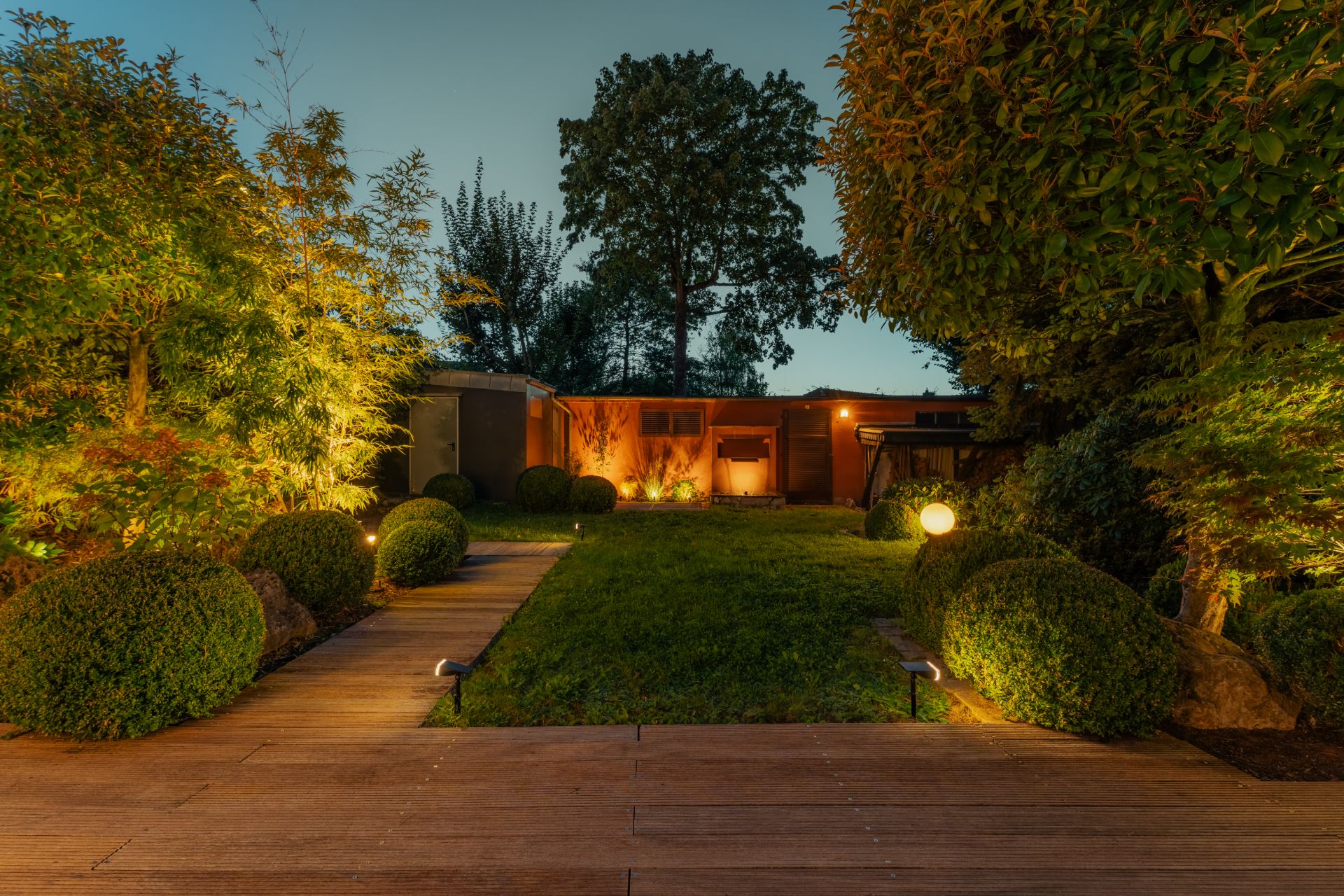 Abendlicher Garten mit beleuchtetem Weg, gepflegtem Rasen und Sträuchern, im Hintergrund ein flaches Haus.