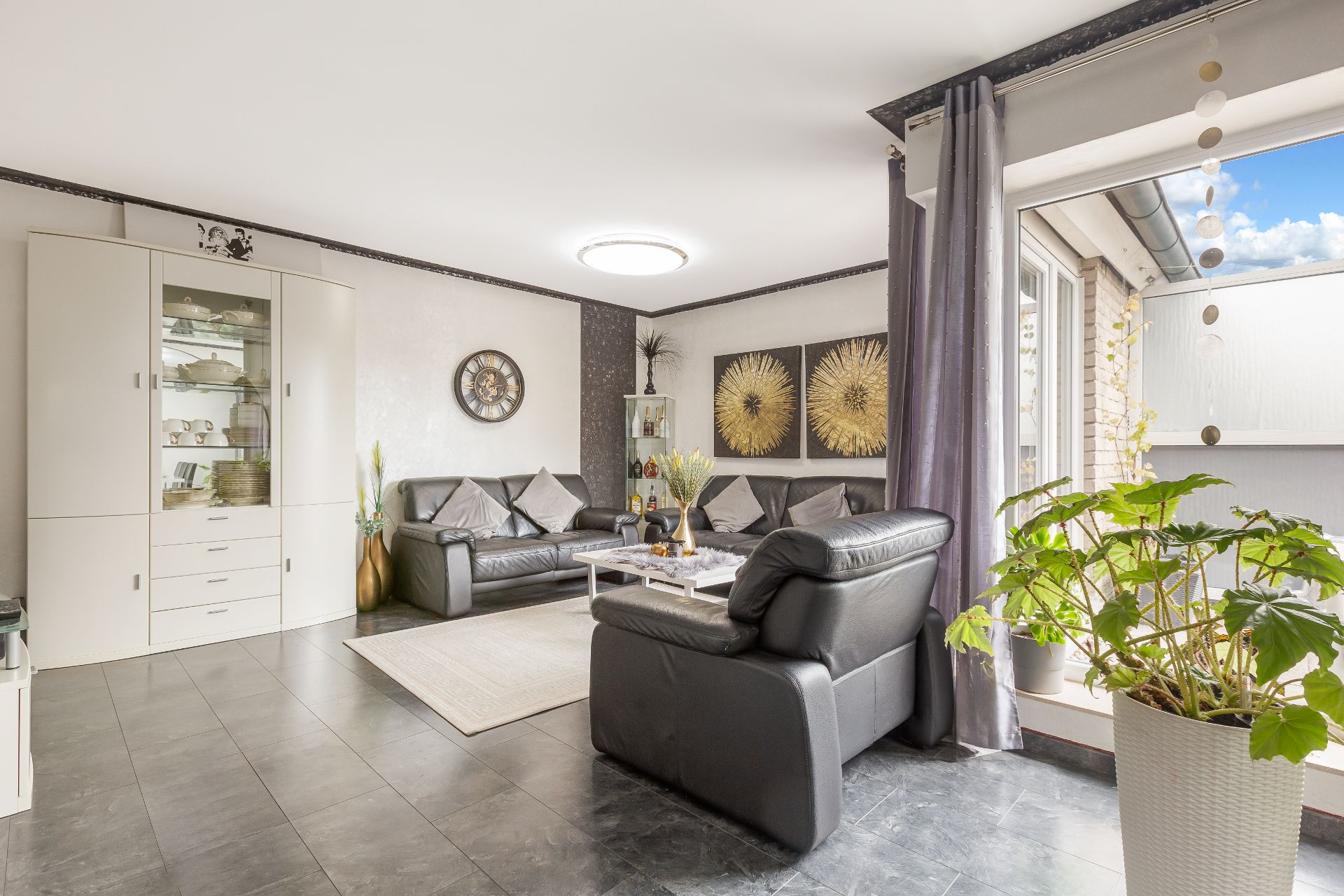 Helles Wohnzimmer mit schwarzen Ledersofas, Vitrine, Teppich und großer Zimmerpflanze am Fenster.