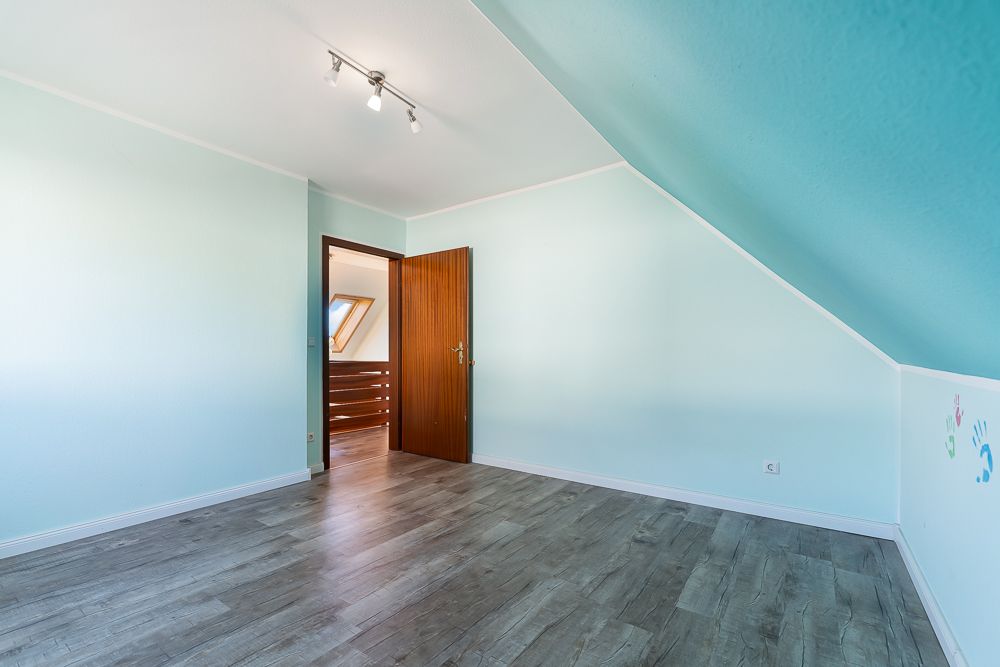 Leeres Zimmer mit hellblauen Wänden, Holztür, Dachschräge und Laminatboden.
