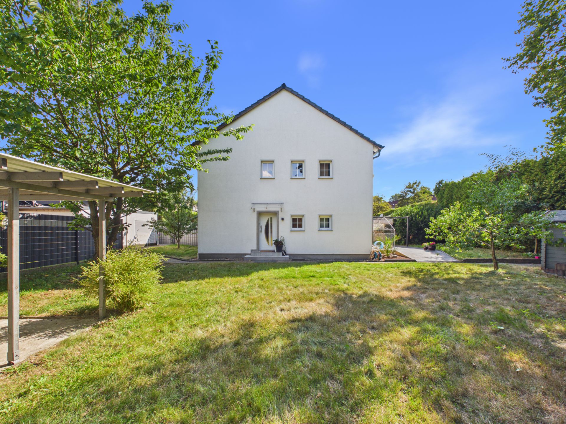 Weißes Einfamilienhaus mit Garten, Bäumen und Rasenfläche an einem sonnigen Tag.