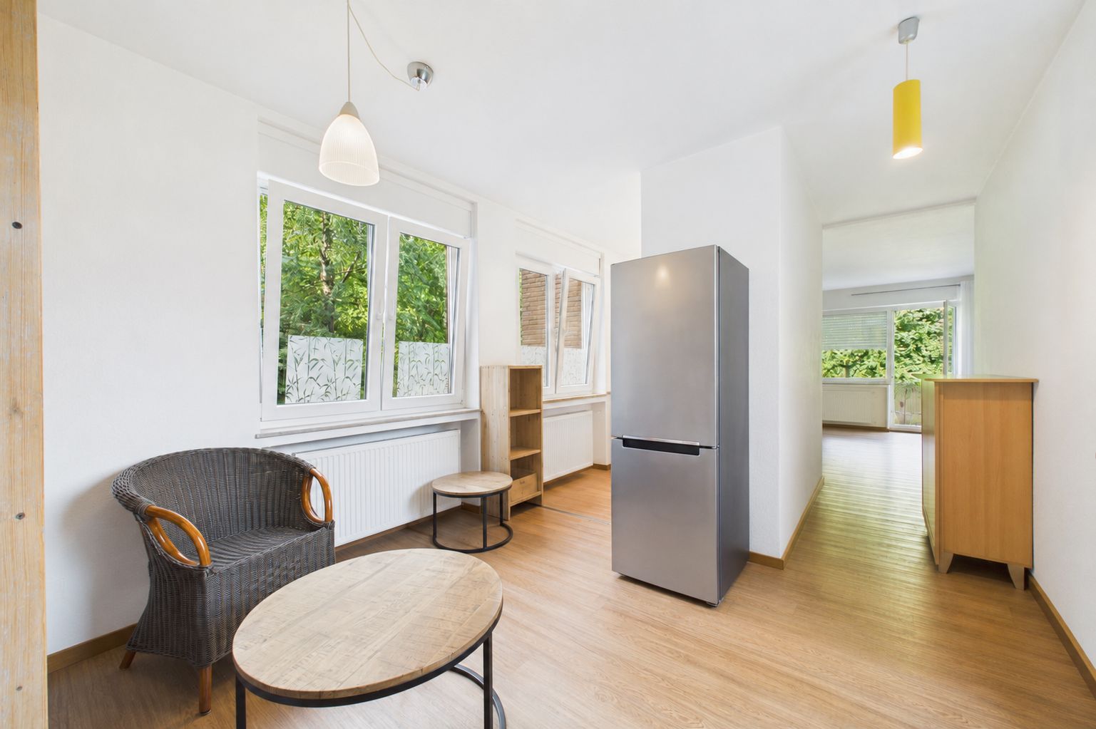 Heller Raum mit Korbstuhl, Holztisch, Kühlschrank und Regalen, große Fenster mit Blick ins Grüne.