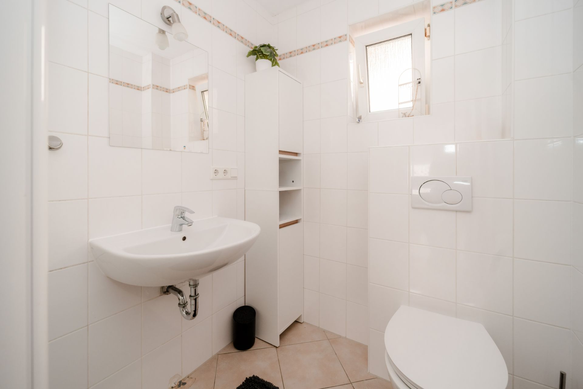 Helles Badezimmer mit Wandwaschbecken, Toilette, Spiegelschrank und kleinem Fenster.