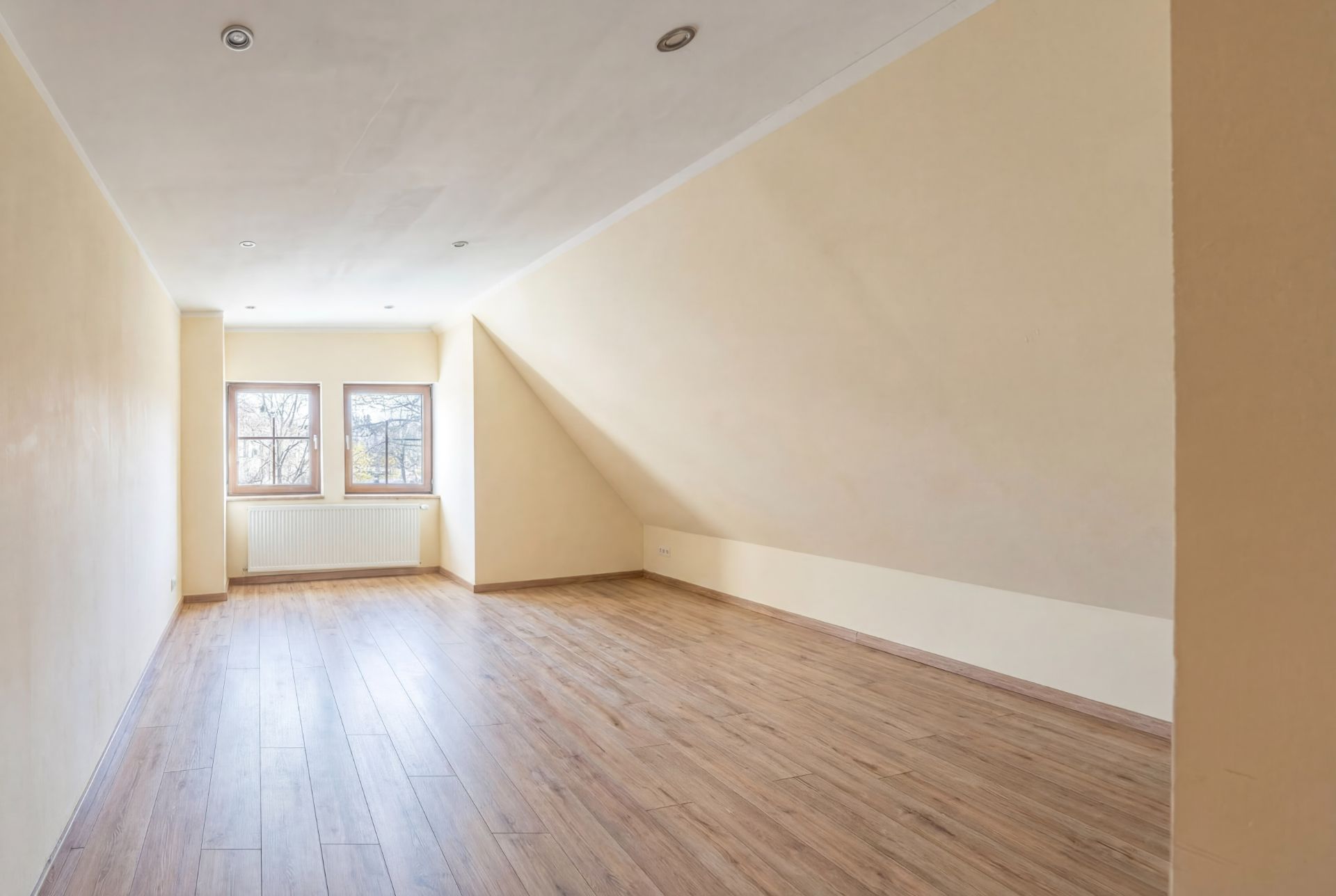 Leerer Raum mit schräger Decke, zwei Fenstern und hellem Holzboden.