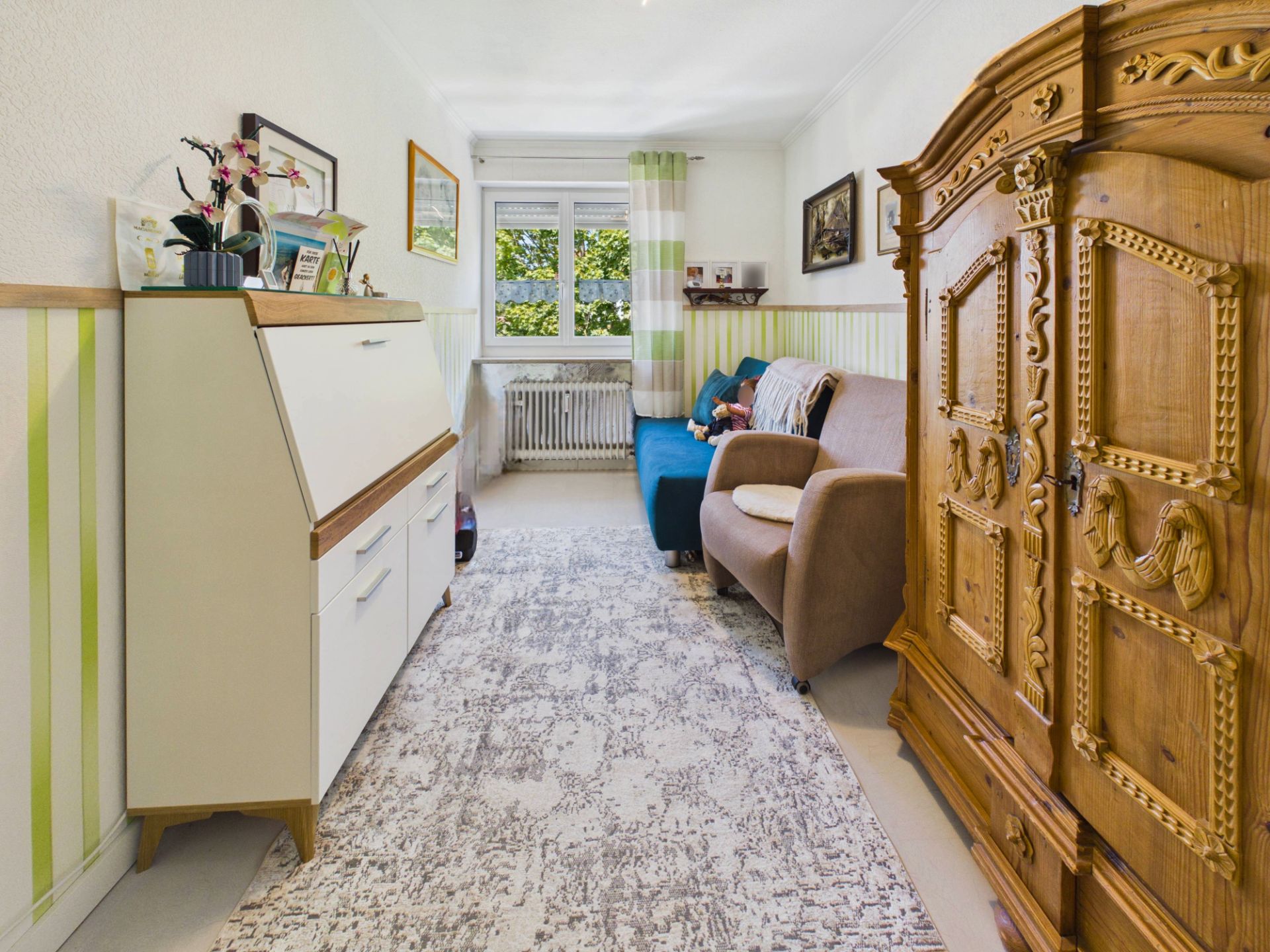 Wohnzimmer mit Sofa, Teppich, großem Holzschrank und Fenster mit Blick ins Grüne.