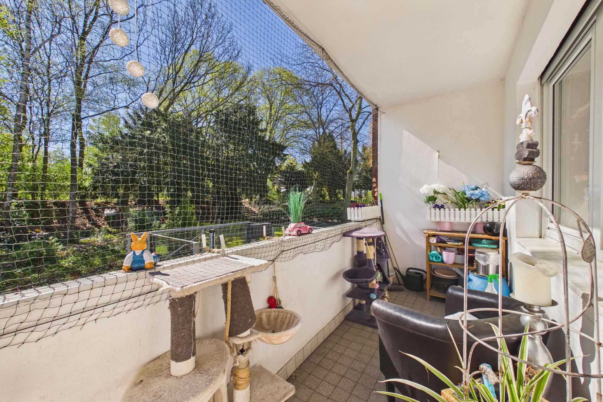 Balkon mit Katzennetz, Kratzbaum, Pflanzen und Blick auf grüne Bäume.