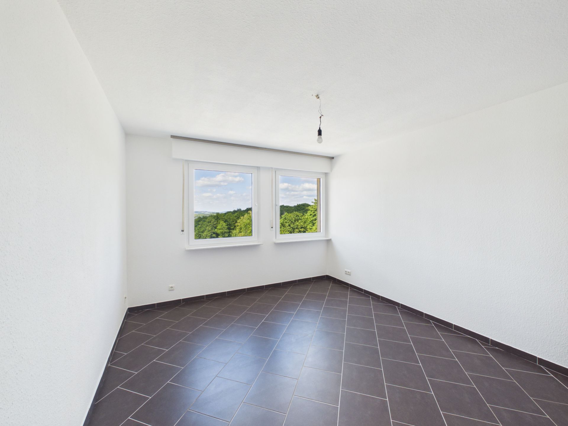 Leeres Zimmer mit weißen Wänden, braunen Fliesen und zwei Fenstern mit Ausblick ins Grüne.