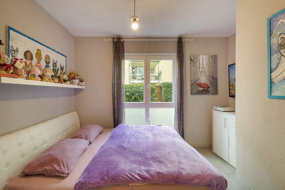 Kleines Schlafzimmer mit Doppelbett, lila Decke, Regal mit Kuscheltieren und großem Fenster.