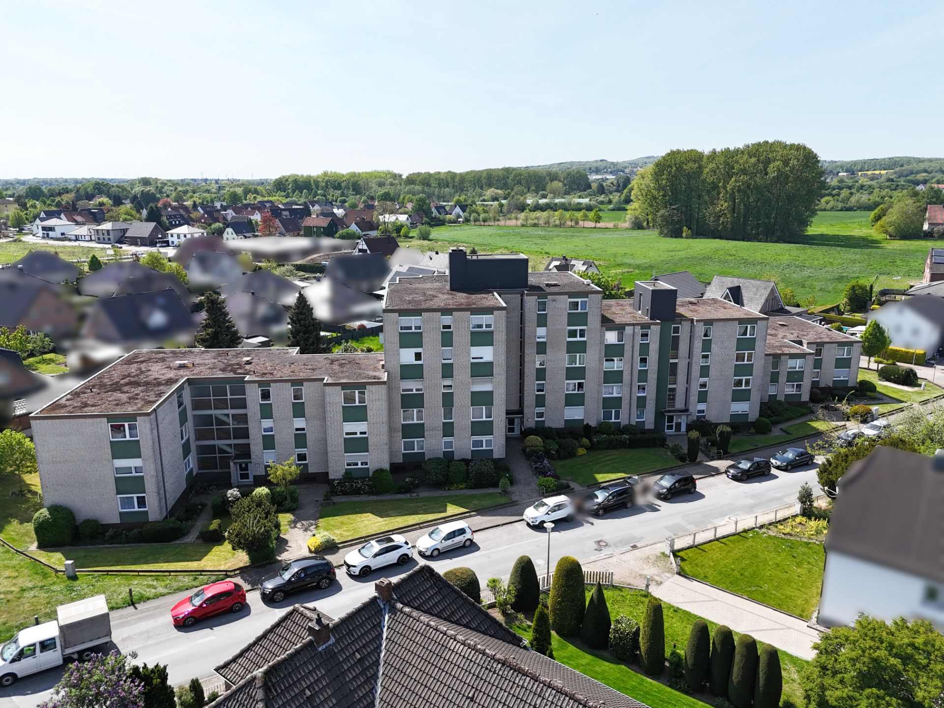 Mehrfamilienhaus mit mehreren Stockwerken und parkenden Autos davor, umgeben von Grünflächen.