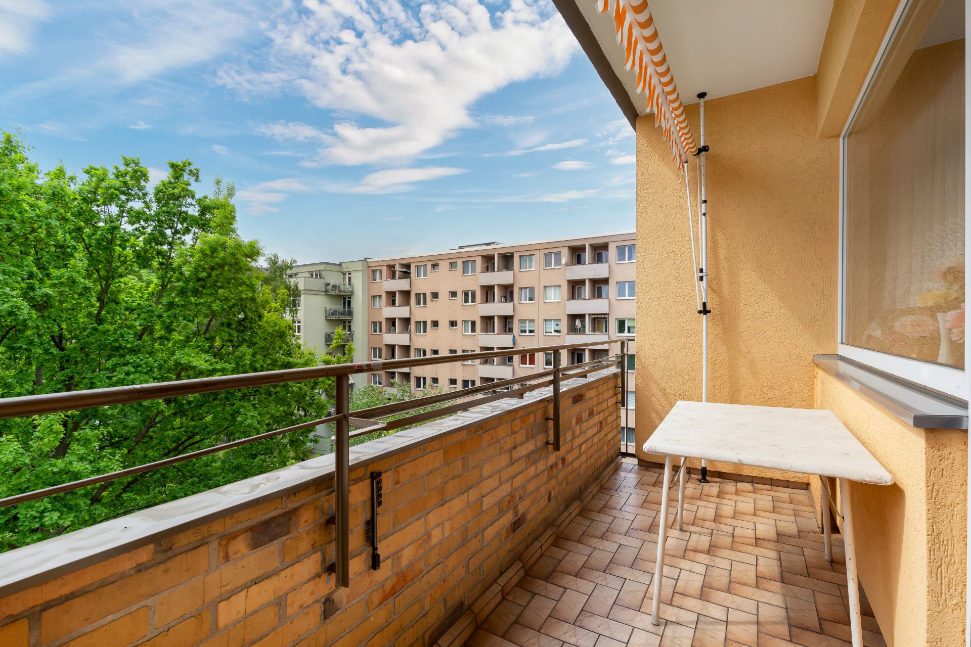 Balkon mit Tisch, Geländer und Markise, Blick auf Bäume und Wohngebäude.