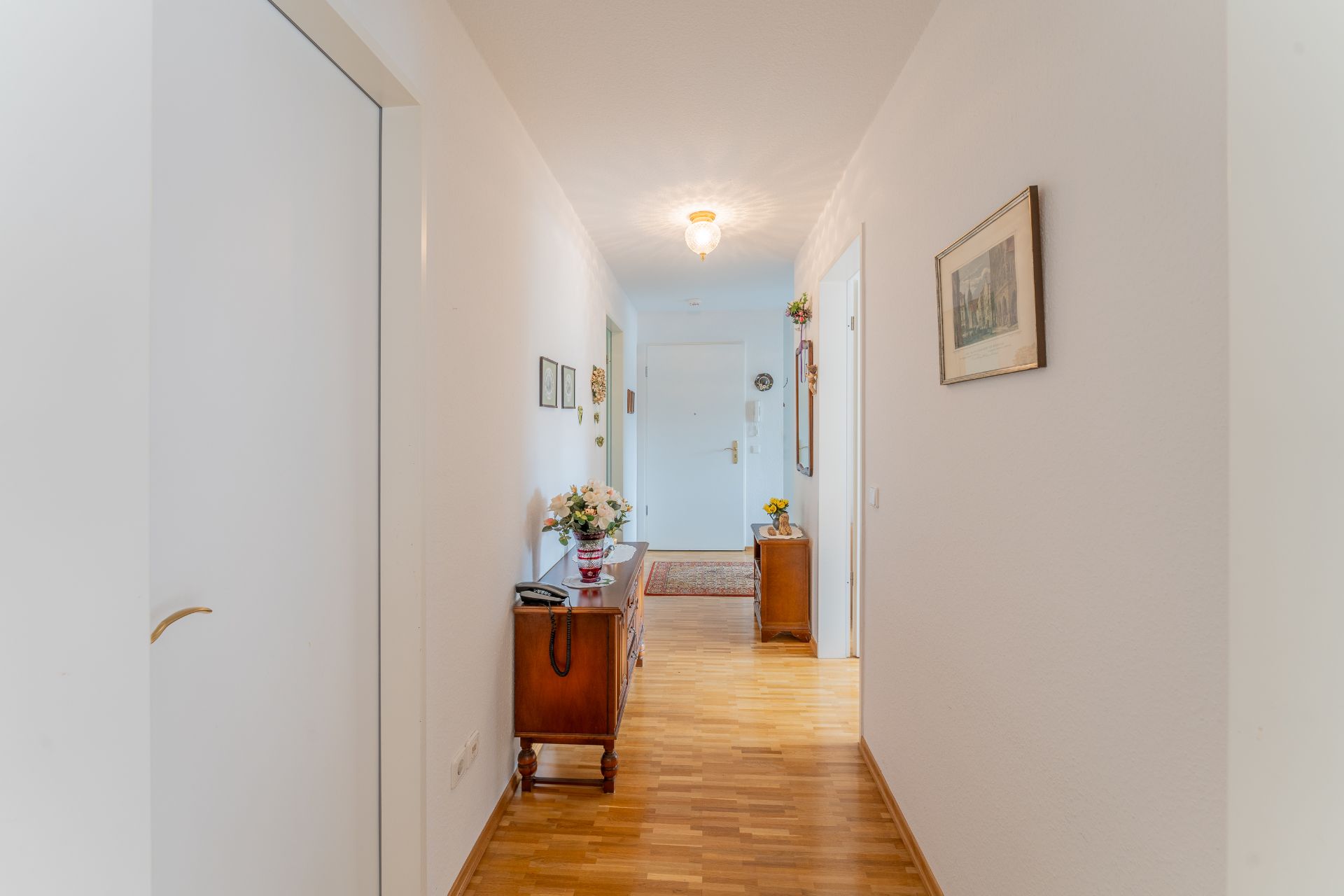 Heller Flur mit Holzboden, Kommoden, Blumenvasen und Bildern an den weißen Wänden.