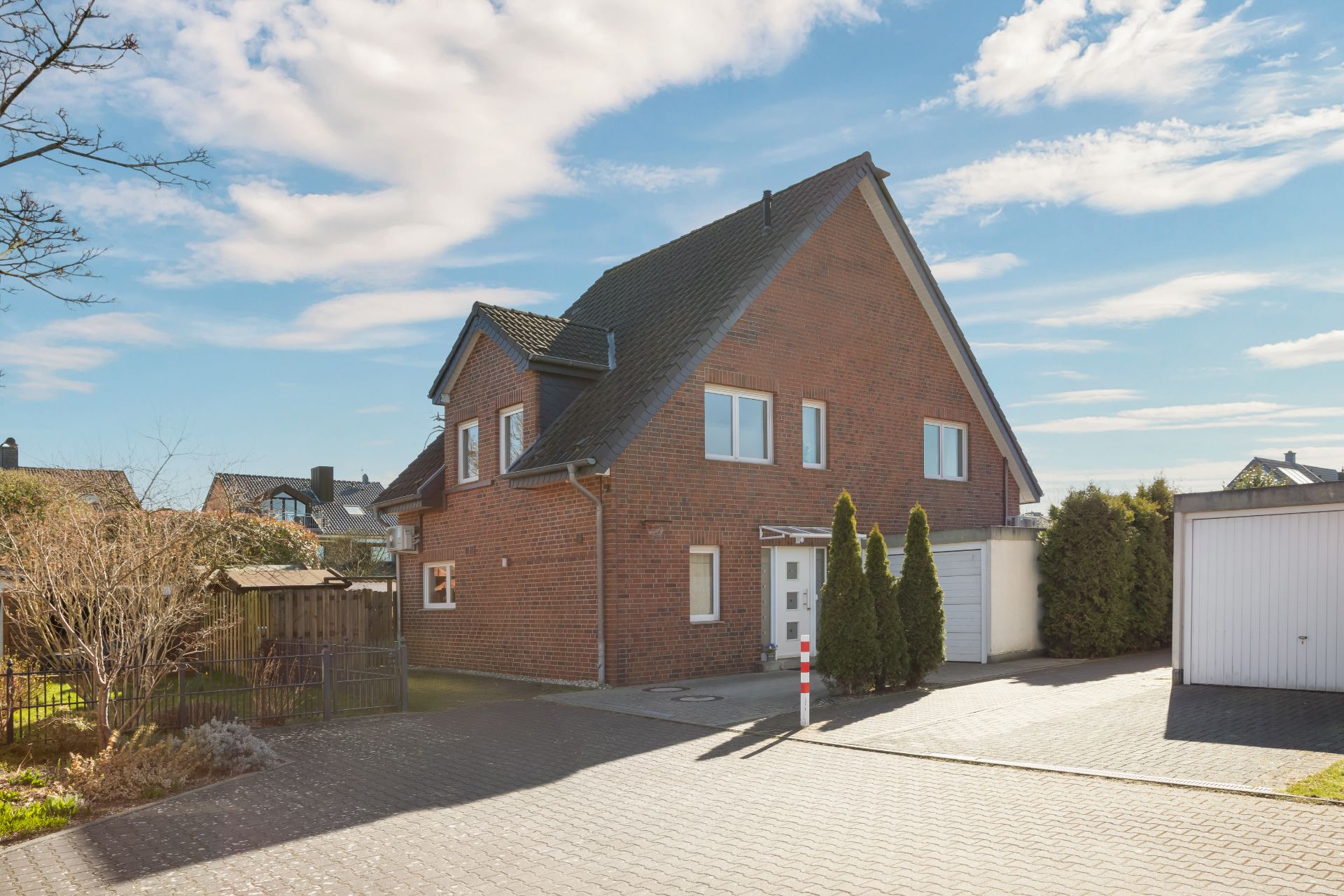 Modernes zweistöckiges Backsteinhaus mit Satteldach, Einfahrt und Garage, umgeben von Hecken und Bäumen.