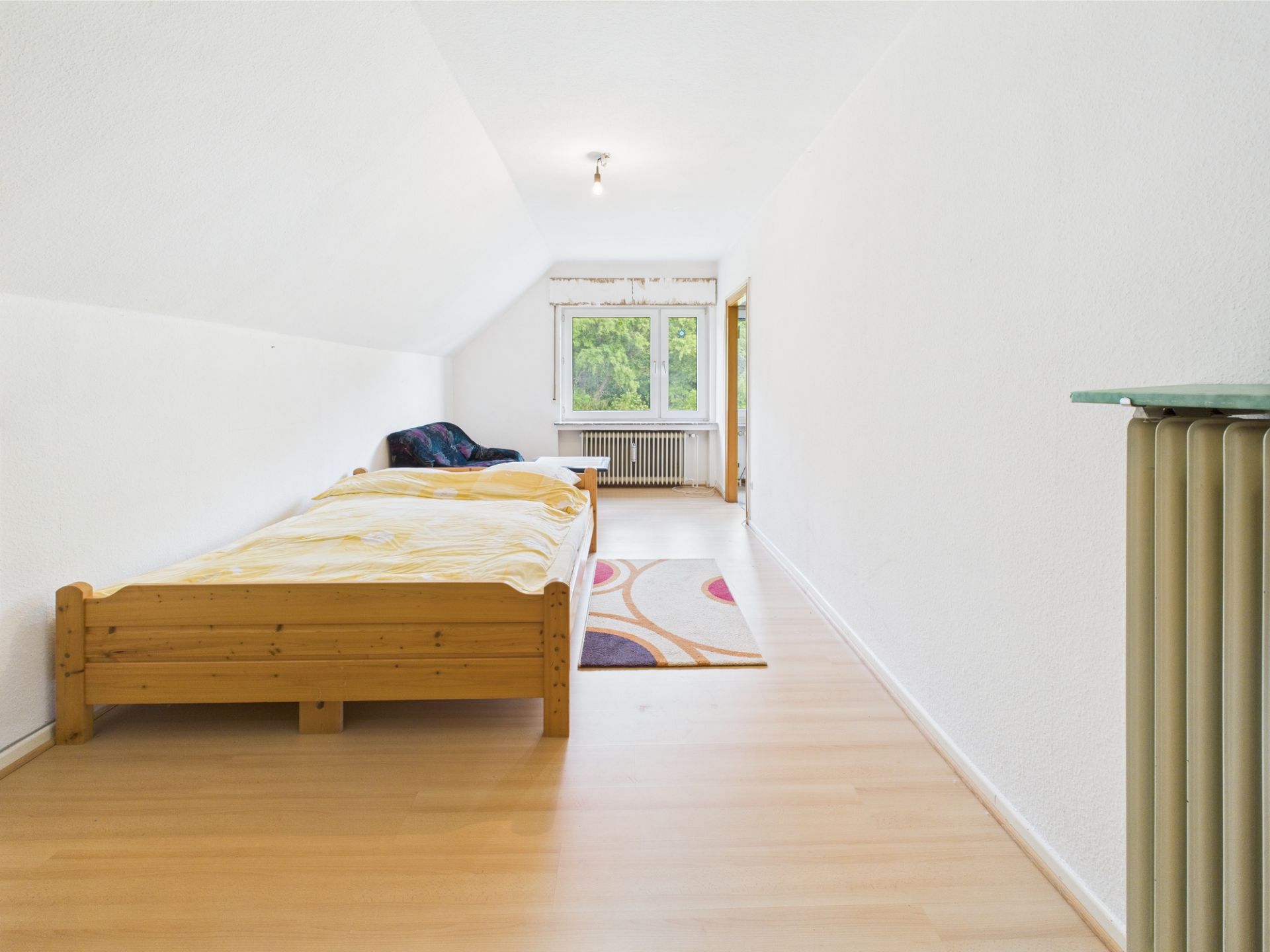 Helles Schlafzimmer mit Holzbett, Teppich und Fenster mit Blick ins Grüne.