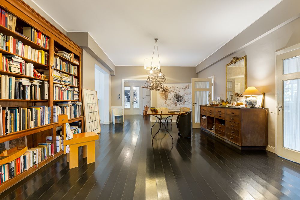 Wohnzimmer mit Bücherregal, Holzstuhl, Kommode, großem Spiegel und Tisch auf dunklem Holzboden.