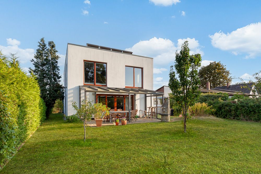 Modernes Einfamilienhaus mit großer Terrasse und Garten, umgeben von Bäumen und Rasen.