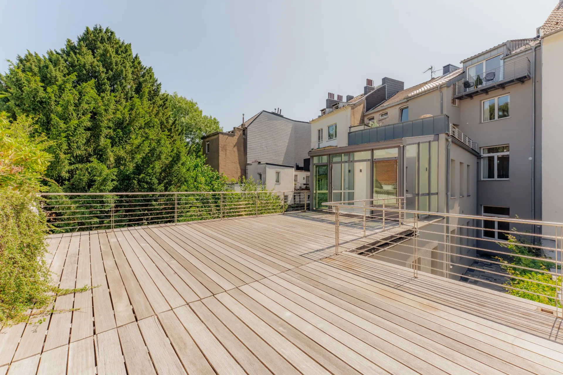 Große Holzterrasse mit Geländer und Blick auf ein modernes Wohnhaus und Bäume.