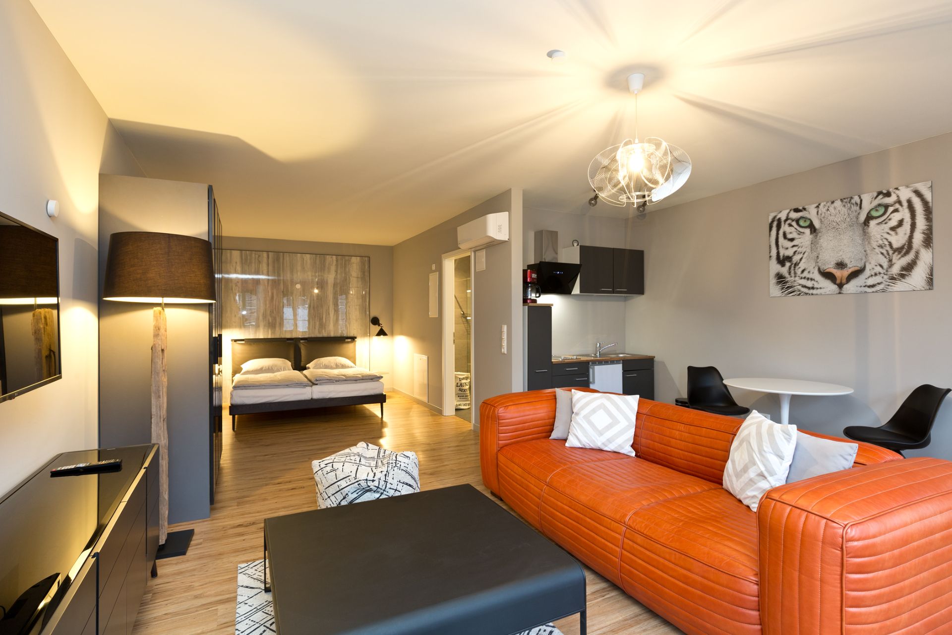 Modern eingerichtetes Apartment mit orangefarbenem Sofa, Bett, Küchenzeile und Tigerbild an der Wand.
