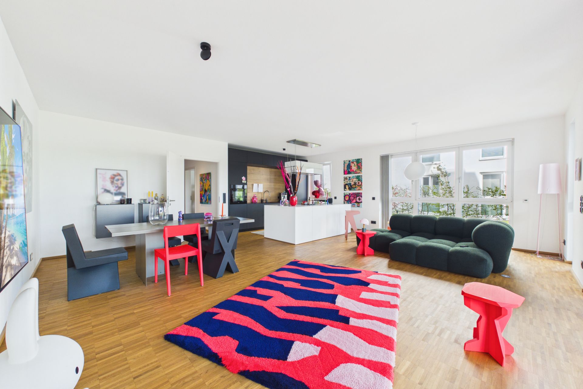 Helles Wohnzimmer mit offener Küche, grünem Sofa, buntem Teppich und Esstisch mit farbigen Stühlen.