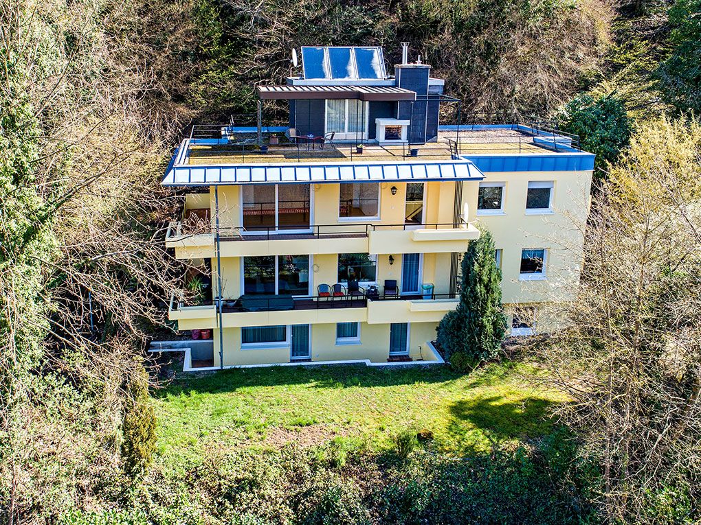 Modernes gelbes Wohnhaus mit Balkonen und Dachterrasse, umgeben von Bäumen.