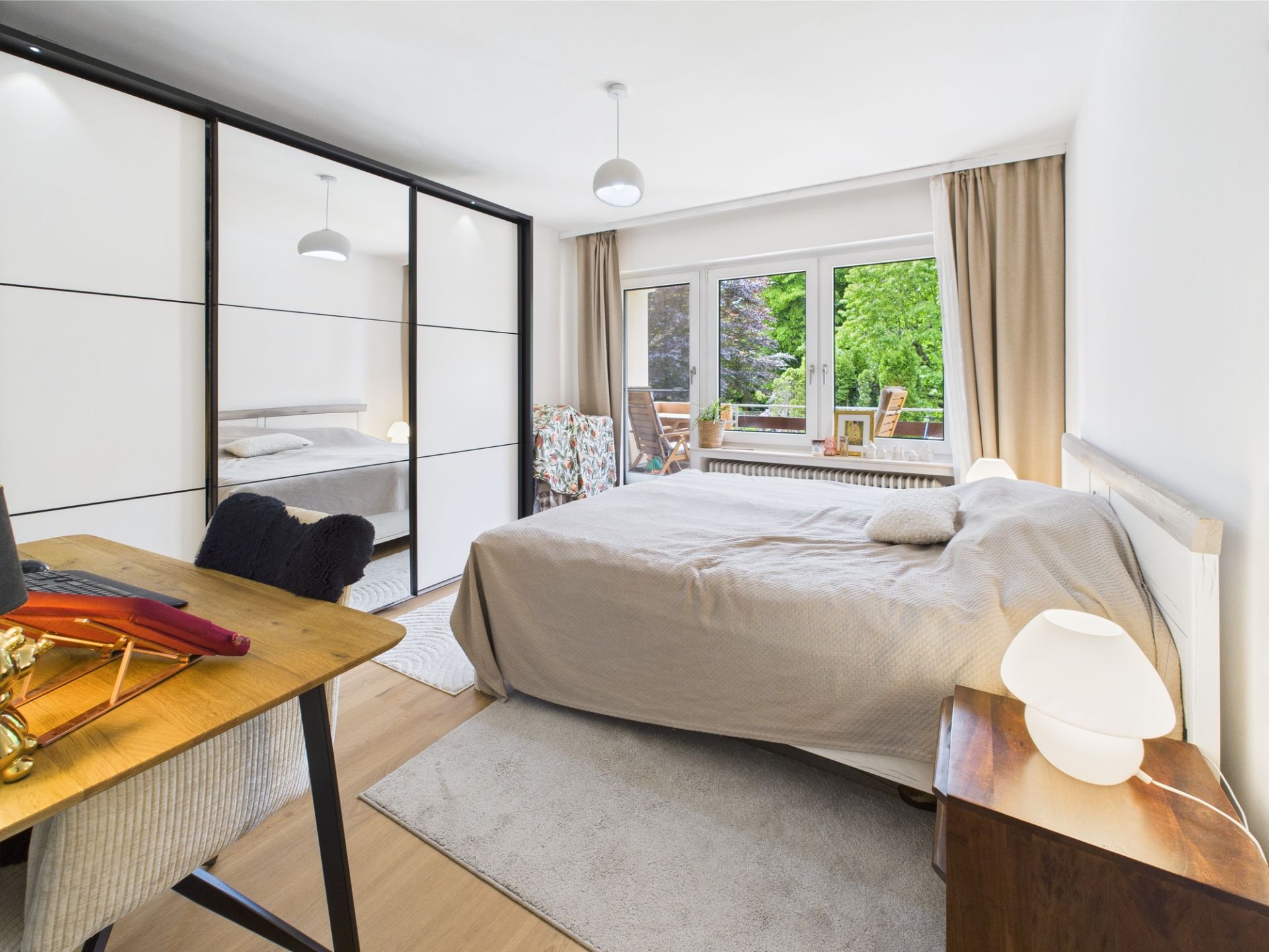 Helles Schlafzimmer mit Doppelbett, Schreibtisch, großem Schrank und Blick auf Balkon mit Grün.