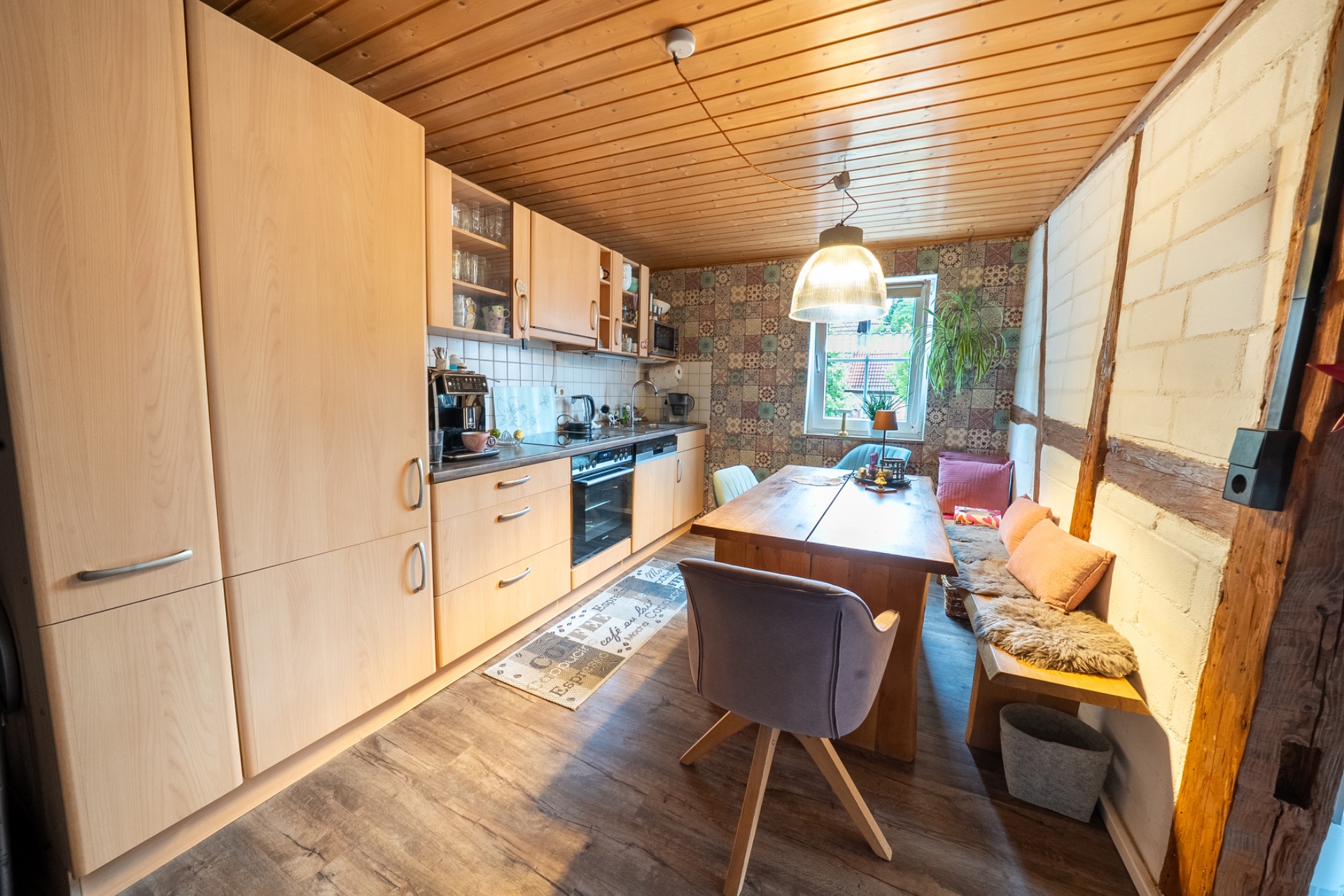 Helle Küche mit Holzdecke, Esstisch, Eckbank, Stühlen und einem Fenster am Ende des Raums.